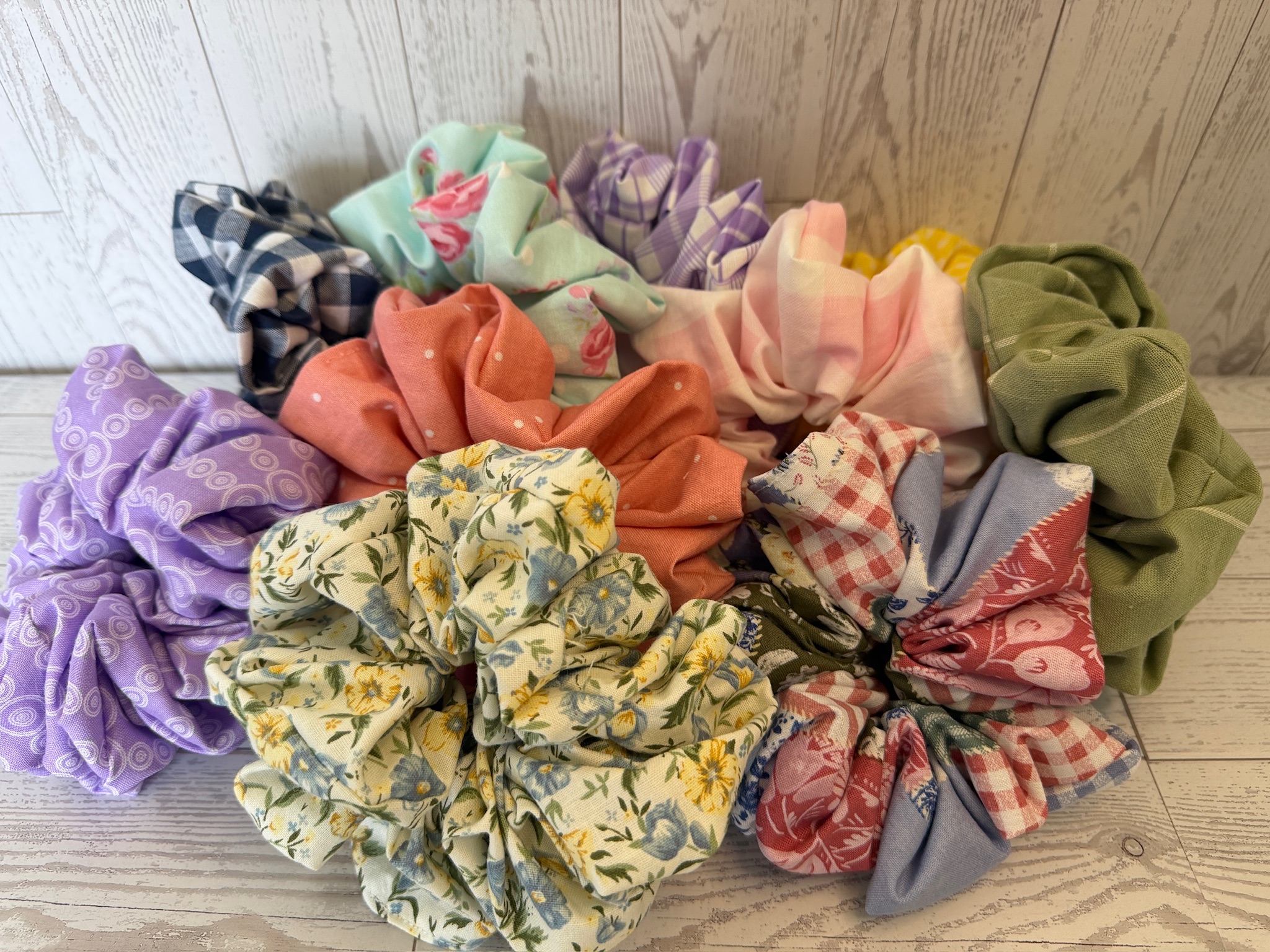 Pile of handmade scrunchies in olive, coral, lavender, buttercup floral, rust gingham and navy plaid fabrics