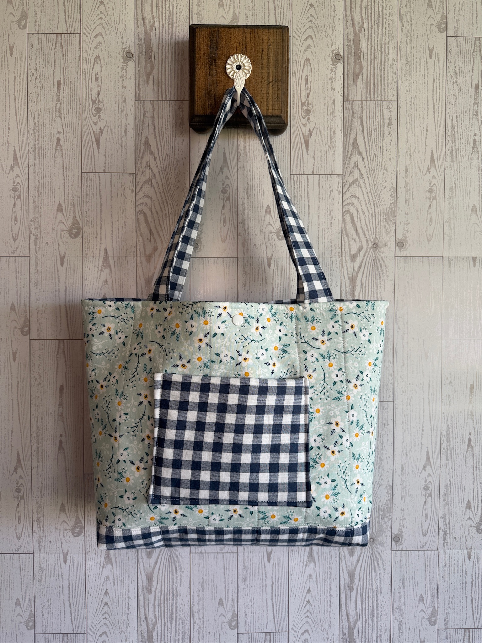 Handmade blue floral tote bag with navy gingham front pocket hanging on a wall hook